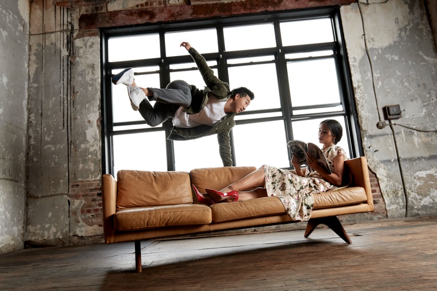 Alex Wong leaping over a sofa while a woman reads beneath him