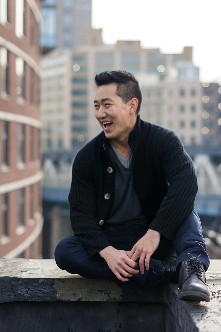 Alex Wong in a black cardigan, laughing on a stone ledge